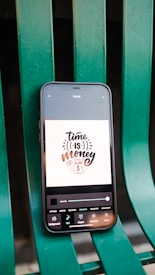 A smartphone displaying a digital art design with the phrase 'time is money' along with icons for currency and a timepiece. The phone is placed on a green metal chair or bench.