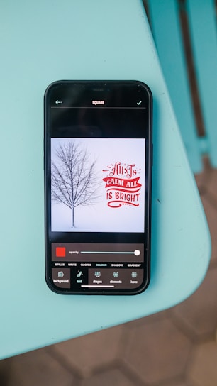 A smartphone lying on a turquoise surface displays an image editing app. The app shows an image of a barren tree against a light background with the text 'All is Calm, All is Bright' in red and white. Various editing tools are visible at the bottom of the screen.