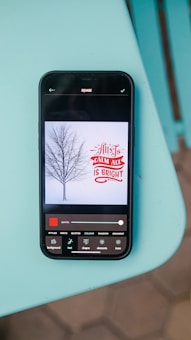 A smartphone lying on a turquoise surface displays an image editing app. The app shows an image of a barren tree against a light background with the text 'All is Calm, All is Bright' in red and white. Various editing tools are visible at the bottom of the screen.