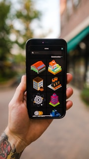A hand holding a smartphone displaying various colorful isometric construction icons. The background is blurred with greenery and a building in an outdoor setting. The phone screen shows icons of buildings, a tower, a factory, an atom symbol, and other related images.