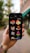 A hand holding a smartphone displaying various colorful isometric construction icons. The background is blurred with greenery and a building in an outdoor setting. The phone screen shows icons of buildings, a tower, a factory, an atom symbol, and other related images.
