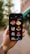 A hand holding a smartphone displaying various colorful isometric construction icons. The background is blurred with greenery and a building in an outdoor setting. The phone screen shows icons of buildings, a tower, a factory, an atom symbol, and other related images.