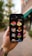 A hand holding a smartphone displaying various colorful isometric construction icons. The background is blurred with greenery and a building in an outdoor setting. The phone screen shows icons of buildings, a tower, a factory, an atom symbol, and other related images.