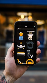 A hand holding a smartphone displaying several fashion and beauty icons. The screen shows images such as a dress, a face with a hat, a comb, a razor, and other beauty-related icons. The background appears out of focus, adding emphasis to the phone's screen.