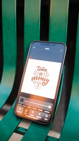 A smartphone displaying an app interface with a design that reads 'Time is Money' alongside a money bag icon. The phone is placed on a green, metal bench with visible slats.