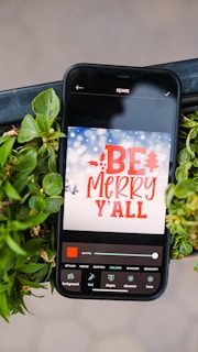 A smartphone displaying a holiday-themed message surrounded by vibrant green leaves. The screen shows a design app with the words 'BE MERRY Y'ALL' in bold red font against a snowy winter background with softly blurred lights. Various editing options are visible at the bottom of the screen.