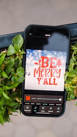 A smartphone displaying a holiday-themed message surrounded by vibrant green leaves. The screen shows a design app with the words 'BE MERRY Y'ALL' in bold red font against a snowy winter background with softly blurred lights. Various editing options are visible at the bottom of the screen.