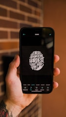 A tattooed hand holding a smartphone displaying a motivational quote, 'First step is the hardest, just make it,' on a dark background. The phone screen shows editing options at the bottom.