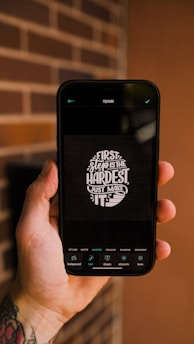 A tattooed hand holding a smartphone displaying a motivational quote, 'First step is the hardest, just make it,' on a dark background. The phone screen shows editing options at the bottom.