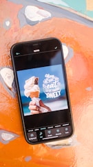A smartphone displaying an image editing application with a photo of a hand holding an ice cream cone. The text on the screen says, 'You are what you eat, so eat something sweet.' The phone is placed on a colorful, abstract background.