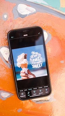 A smartphone displaying an image editing application with a photo of a hand holding an ice cream cone. The text on the screen says, 'You are what you eat, so eat something sweet.' The phone is placed on a colorful, abstract background.