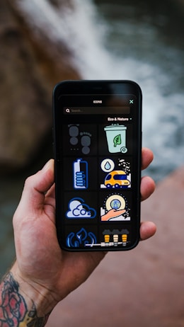 A tattooed hand holds a smartphone displaying a set of eco and nature-themed icons against a blurred natural background. The icons include a recycling bin, cloud with CO2, a green energy symbol, among others.