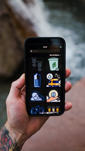 A tattooed hand holds a smartphone displaying a set of eco and nature-themed icons against a blurred natural background. The icons include a recycling bin, cloud with CO2, a green energy symbol, among others.