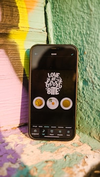 A smartphone rests against a colorfully painted wooden surface. The phone screen displays the phrase 'Love at first bite' along with images of food items such as noodles, an orange, and a cup of coffee. The wooden surface features a blend of vibrant colors including green, yellow, pink, and purple.
