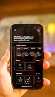 A hand is holding a smartphone displaying a screen with options for selecting the perfect layout for multiple platforms. The smartphone screen shows various platform options such as Instagram, Facebook, TikTok, and more. The background is blurred, focusing attention on the phone.