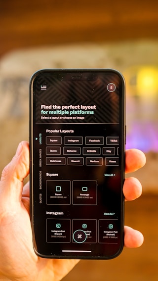 A hand is holding a smartphone displaying a screen with options for selecting the perfect layout for multiple platforms. The smartphone screen shows various platform options such as Instagram, Facebook, TikTok, and more. The background is blurred, focusing attention on the phone.