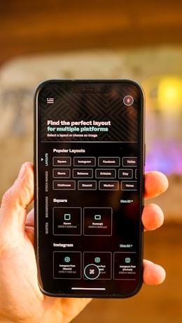 A hand is holding a smartphone displaying a screen with options for selecting the perfect layout for multiple platforms. The smartphone screen shows various platform options such as Instagram, Facebook, TikTok, and more. The background is blurred, focusing attention on the phone.