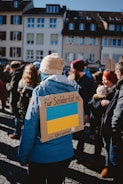 A group of people gathered outdoors, many wearing winter clothing. The central figure wears a blue jacket and a beige knit hat, and has a cardboard sign on their back with the Ukrainian flag and text. The event seems to be a solidarity gathering in support of Ukraine, set in a European urban environment with traditional buildings in the background.
