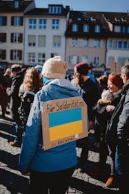 A group of people gathered outdoors, many wearing winter clothing. The central figure wears a blue jacket and a beige knit hat, and has a cardboard sign on their back with the Ukrainian flag and text. The event seems to be a solidarity gathering in support of Ukraine, set in a European urban environment with traditional buildings in the background.