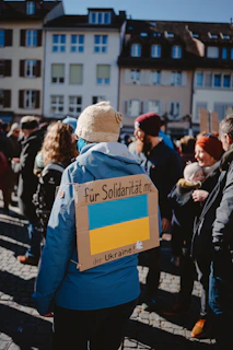 A group of people gathered outdoors, many wearing winter clothing. The central figure wears a blue jacket and a beige knit hat, and has a cardboard sign on their back with the Ukrainian flag and text. The event seems to be a solidarity gathering in support of Ukraine, set in a European urban environment with traditional buildings in the background.
