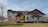 A modern two-story house with a mix of wood and stone elements, surrounded by a sparse landscape with a few leafless trees. The house features large windows, a spacious front patio with chairs, and a prominent wooden gable. The sky is clear with some clouds, and there is a water body visible in the distance.