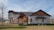 A modern two-story house with a mix of wood and stone elements, surrounded by a sparse landscape with a few leafless trees. The house features large windows, a spacious front patio with chairs, and a prominent wooden gable. The sky is clear with some clouds, and there is a water body visible in the distance.