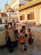 A group of young boys dressed in traditional robes engage in water play near a decorative and ornate building reminiscent of a temple. The setting is lively and captures a moment of playful interaction as they use buckets of water, with the architecture in the background displaying intricate golden accents.
