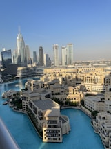 A stunning cityscape featuring modern skyscrapers and a luxurious waterfront development. The skyline includes tall, gleaming towers, while a large, tranquil body of turquoise water surrounds ornate buildings with traditional architectural elements. The setting is bathed in natural daylight, suggesting a vibrant, lively atmosphere.