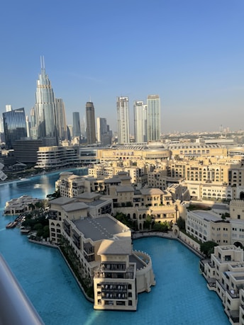 A stunning cityscape featuring modern skyscrapers and a luxurious waterfront development. The skyline includes tall, gleaming towers, while a large, tranquil body of turquoise water surrounds ornate buildings with traditional architectural elements. The setting is bathed in natural daylight, suggesting a vibrant, lively atmosphere.