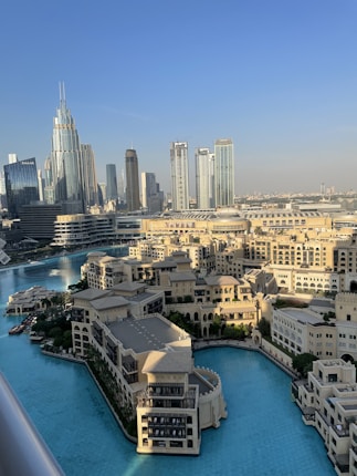 A stunning cityscape featuring modern skyscrapers and a luxurious waterfront development. The skyline includes tall, gleaming towers, while a large, tranquil body of turquoise water surrounds ornate buildings with traditional architectural elements. The setting is bathed in natural daylight, suggesting a vibrant, lively atmosphere.