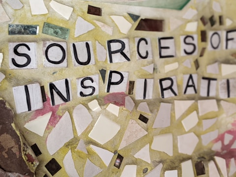 A mosaic of scattered, irregularly shaped tiles in various shades of yellow, white, and pastel colors. Bold black letters spell out the phrase 'SOURCES OF INSPIRATION' on the tiles. The arrangement creates a textured and visually engaging composition.