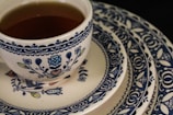 A ceramic teacup filled with tea rests on a stack of ornately patterned plates. The design features intricate blue floral and geometric motifs against a cream-colored background.
