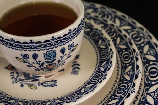 A ceramic teacup filled with tea rests on a stack of ornately patterned plates. The design features intricate blue floral and geometric motifs against a cream-colored background.