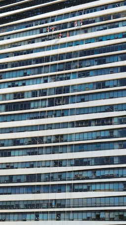 A group of workers in red uniforms are suspended by ropes and cleaning the windows of a tall office building. The building has multiple floors with a repetitive pattern of glass windows and white horizontal bands.
