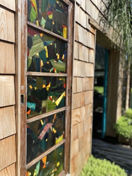 A wooden exterior wall with a vertical stained glass window panel. The window features a mosaic design with various colors including green, red, and yellow. Sunlight highlights the wood grain and the colorful reflections on the glass. In the background, green foliage and part of another opening are visible.