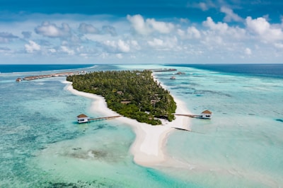 A lush green island surrounded by clear turquoise waters and white sandy beaches. Wooden piers extend from the island into the shallow waters. Overwater bungalows are visible in the distance, built on stilts above the sea. The sky is partly cloudy with patches of blue, adding to the picturesque tropical setting.
