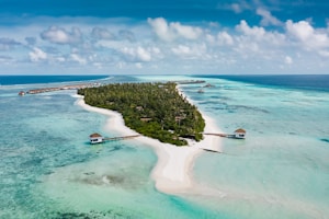 A lush green island surrounded by clear turquoise waters and white sandy beaches. Wooden piers extend from the island into the shallow waters. Overwater bungalows are visible in the distance, built on stilts above the sea. The sky is partly cloudy with patches of blue, adding to the picturesque tropical setting.