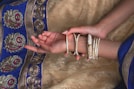 A pair of hands adorned with multiple gold bangles rests on a richly decorated fabric with intricate patterns. The fabric features detailed embroidery with gold, blue, and magenta colors.