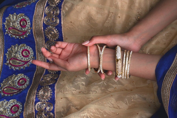 A pair of hands adorned with multiple gold bangles rests on a richly decorated fabric with intricate patterns. The fabric features detailed embroidery with gold, blue, and magenta colors.