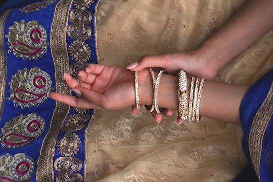 A pair of hands adorned with multiple gold bangles rests on a richly decorated fabric with intricate patterns. The fabric features detailed embroidery with gold, blue, and magenta colors.