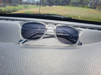 A pair of clear-framed glasses with dark lenses is placed on a car dashboard. The surface of the dashboard has a textured, leather-like appearance, and the background shows a blurred view of the outdoor scenery, including grass and a few buildings.