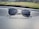 A pair of clear-framed glasses with dark lenses is placed on a car dashboard. The surface of the dashboard has a textured, leather-like appearance, and the background shows a blurred view of the outdoor scenery, including grass and a few buildings.