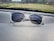 A pair of clear-framed glasses with dark lenses is placed on a car dashboard. The surface of the dashboard has a textured, leather-like appearance, and the background shows a blurred view of the outdoor scenery, including grass and a few buildings.