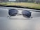 A pair of clear-framed glasses with dark lenses is placed on a car dashboard. The surface of the dashboard has a textured, leather-like appearance, and the background shows a blurred view of the outdoor scenery, including grass and a few buildings.
