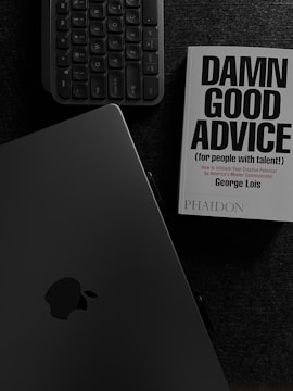A book with the title 'Damn Good Advice (for people with talent!)' by George Lois is placed on a dark surface alongside a keyboard and an open laptop displaying the Apple logo. The overall color scheme is monochromatic, dominated by dark shades, enhancing a professional ambiance.