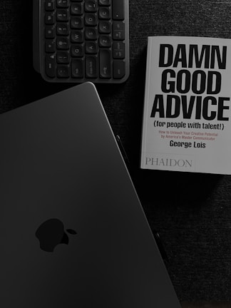 A book with the title 'Damn Good Advice (for people with talent!)' by George Lois is placed on a dark surface alongside a keyboard and an open laptop displaying the Apple logo. The overall color scheme is monochromatic, dominated by dark shades, enhancing a professional ambiance.