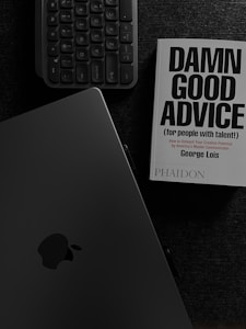 A book with the title 'Damn Good Advice (for people with talent!)' by George Lois is placed on a dark surface alongside a keyboard and an open laptop displaying the Apple logo. The overall color scheme is monochromatic, dominated by dark shades, enhancing a professional ambiance.