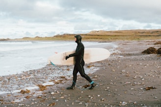A surfer in a black wetsuit carrying a white surfboard walks along a pebble-strewn beach. The sky is overcast with clouds, and waves are gently crashing on the shoreline. The backdrop features rolling hills and a grassy landscape, creating a serene coastal environment.
