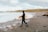 A surfer in a black wetsuit carrying a white surfboard walks along a pebble-strewn beach. The sky is overcast with clouds, and waves are gently crashing on the shoreline. The backdrop features rolling hills and a grassy landscape, creating a serene coastal environment.