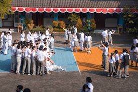 A large group of students in school uniforms, mostly in white shirts, are gathered outdoors in a school courtyard. Some are taking selfies while standing on colorfully painted sections of the ground. Others are mingling, and some are sitting on the ground. The building in the background has a tiled roof and is decorated with red and white bunting.